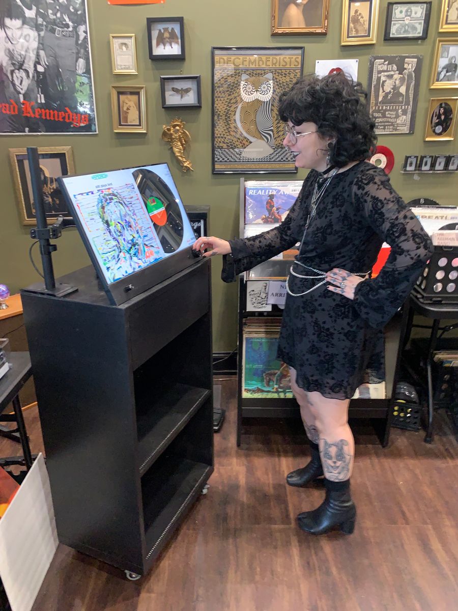 A shop owner turning the control knob on the Almont display in a record store. The tilted touchscreen shows a life-size album cover beside its vinyl record and label.